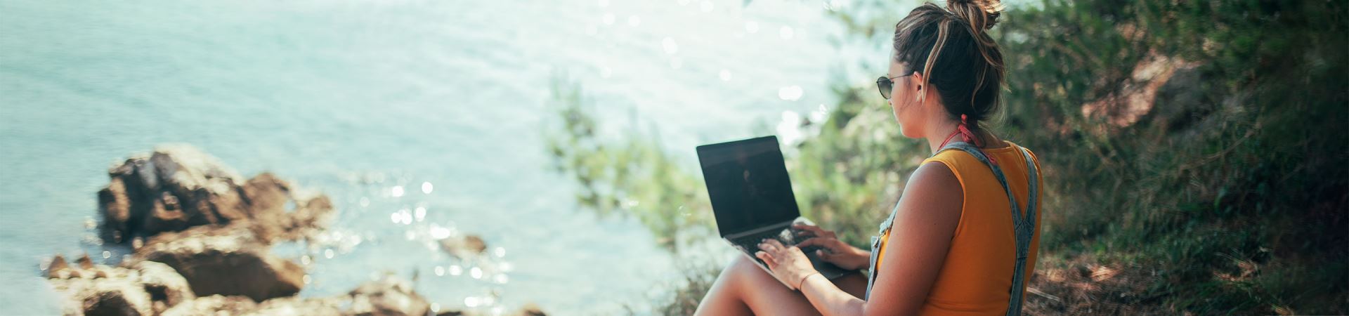 Das Bild zeigt ein Mädchen im Profil mit einem Laptop auf dem Schoß. Es trägt einen orangefarbenen Pullover und einen Jeansoverall und sitzt in einem Waldgebiet mit Blick auf das Meer.