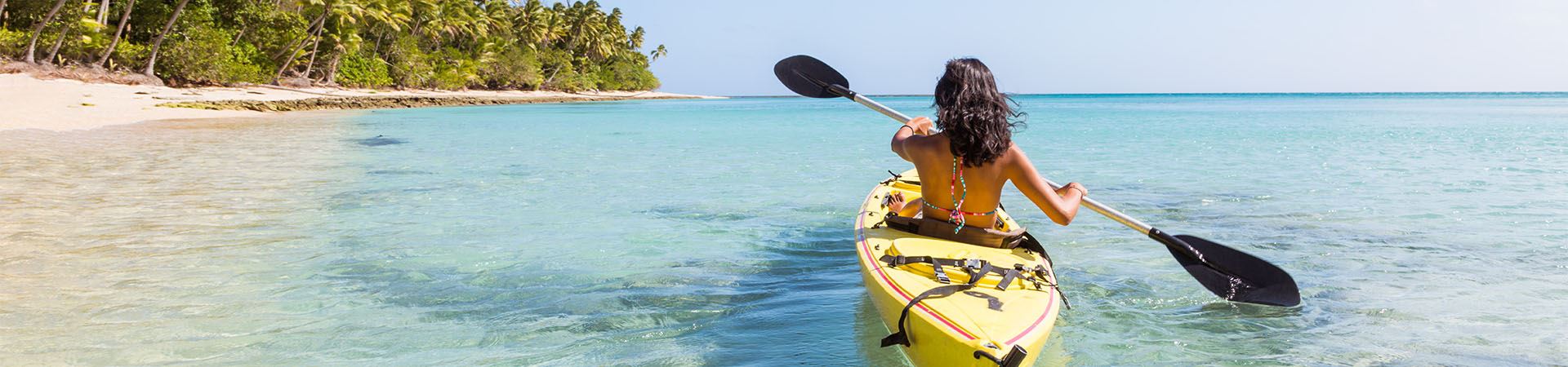 This picture shows a girl riding a canoe in the sea, seen from behind. The canoe is yellow and the background consists of sand and some vegetation, suggesting it is close to the beach.