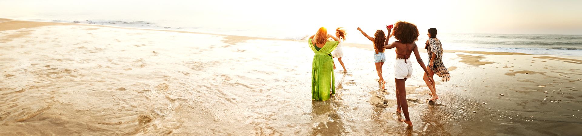 Cette image montre cinq filles au bord de la mer. L’une d’elles a les bras en l’air et une autre court. Elles portent toutes des vêtements légers et d’été. En arrière-plan, on voit la mer et la lumière indique que c'est le coucher du soleil.