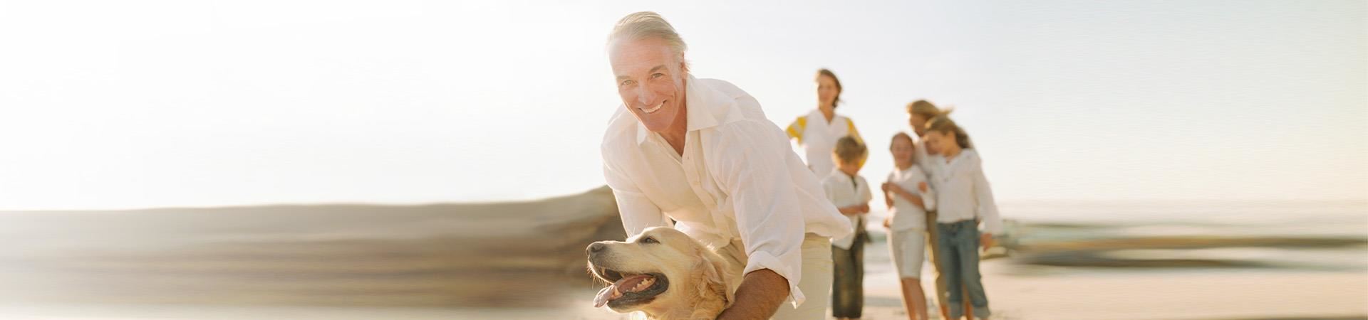Image of a gray-haired man with blue eyes on the beach, leaning forward while holding a beige golden retriever. The man is wearing a white shirt and beige pants. Slightly blurred in the background to his right, we can see (from left to right) a woman, a boy, two girls, and a teenage girl. 