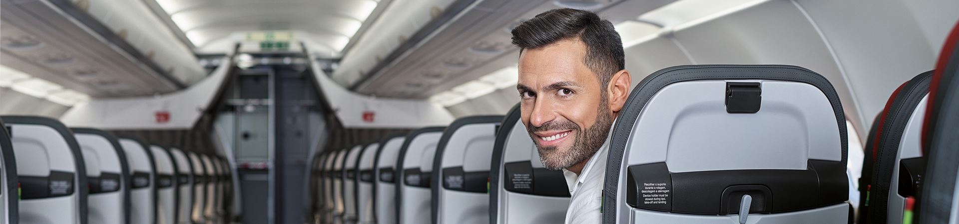 Plan d'un couloir et de l'arrière des sièges du côté droit d'un avion TAP. Dans le dernier siège visible, un homme caucasien aux cheveux noirs est assis, vêtu d'une chemise blanche et regardant en arrière, directement vers la caméra, en souriant. 