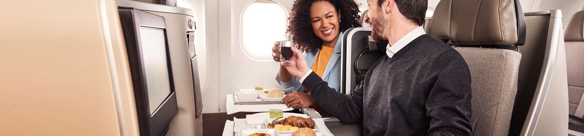 Un homme et une femme, souriants, assis côte à côte à bord d'un avion TAP. Ils tiennent, tous les deux, un verre à la main et portent un toast. Devant eux, chacun a un plateau avec un repas de viande, du pain et un pastel de nata.