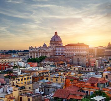 Immagine aerea della città di Roma. In primo piano abbiamo vari edifici residenziali e sullo sfondo vari monumenti della città di Roma con dietro il tramonto.