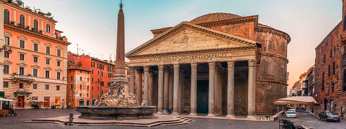 Immagine, in prospettiva laterale, di una via di Roma in cui vediamo in fondo l'edificio del Pantheon di Roma e una fontana. Le vie laterali nell'immagine sono fiancheggiate da edifici color arancio e da auto parcheggiate sulle strade lastricate.