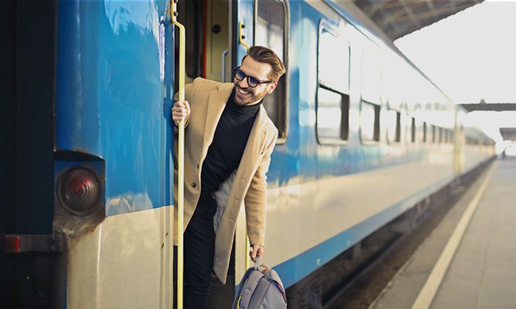 Fotografía de un hombre saliendo por la puerta de un tren azul. El hombre sonríe y sujeta la barandilla de la puerta con la mano derecha, mientras lleva su mochila en la mano izquierda. Está vestido de negro con un abrigo beige.