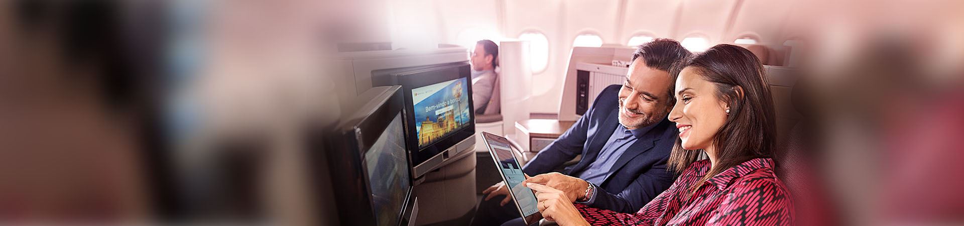 Primer plano de una mujer y un hombre sonrientes y sentados a bordo de un avión. La mujer sostiene una tableta y el hombre la mira. Ambos apuntan a la pantalla de la tableta. En frente, hay dos pantallas integradas en los asientos del avión.