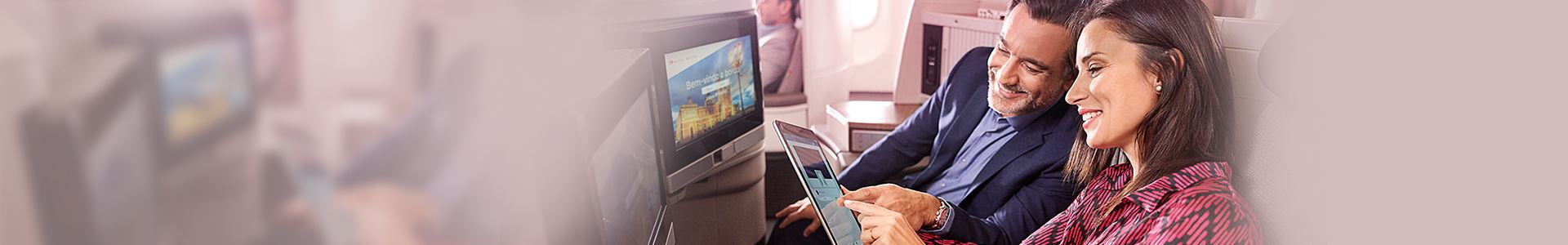 Nahaufnahme einer lächelnden Frau und eines lächelnden Mannes, die in einem Flugzeug sitzen. Die Frau hält ein Tablet in der Hand, und der Mann blickt darauf. Sie zeigen beide auf den Bildschirm des Tablets. Vor ihnen befinden sich zwei Bildschirme, die in den Sitzen des Flugzeugs eingebaut sind.