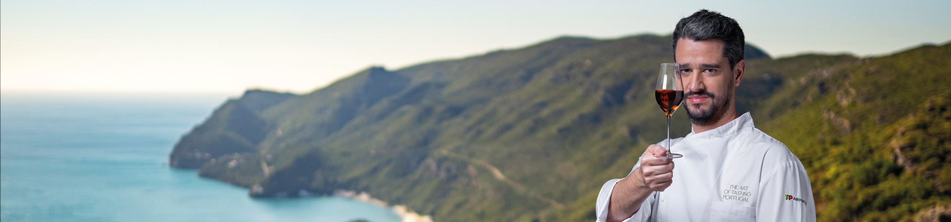 Bild mit den grünen Bergen der Halbinsel Setúbal im Hintergrund. Im Vordergrund, auf der rechten Seite des Bildes, hält der Chefkoch André Cruz ein Glas Moscatel de Setúbal mit der rechten Hand auf Gesichtshöhe und bedeckt dabei teilweise seine rechte Gesichtshälfte.