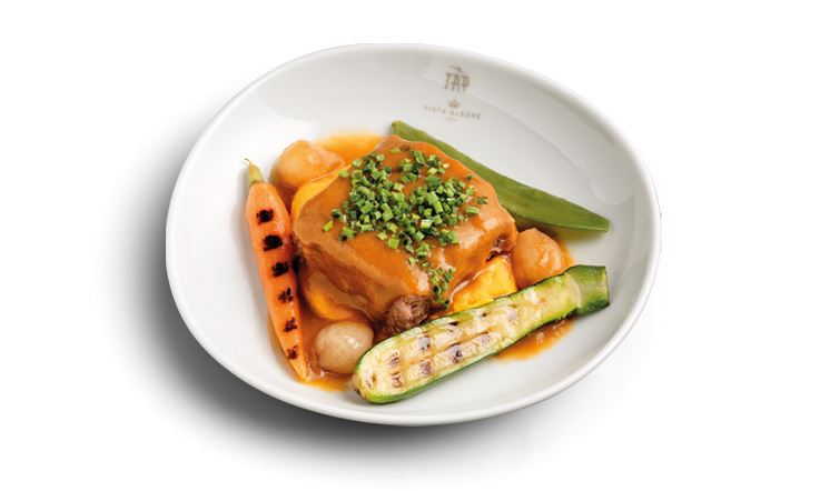 Фотография глубокой белой тарелки с золотистым логотипом TAP Air Portugal на ободке, в центре которой лежит кусок телятины Оссо Буко, покрытый оранжевым соусом Москатель де Сетубал, морковным пюре и весенними овощами.