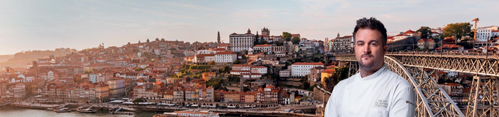 Medium Shot – KochArnaldo Azevedo Er trägt eine weiße Kochschürze mit der Aufschrift „The Art of Tasting Portugal“ an der oberen linken Brusttasche und dem Logo von TAP Air Portugal am linken Ärmel. Der Koch steht vor dem Hintergrund eines Panoramafotos von Porto mit dem Fluss Douro und der Stadt sowie "D. Maria Pia Bridge" rechts zu sehen.