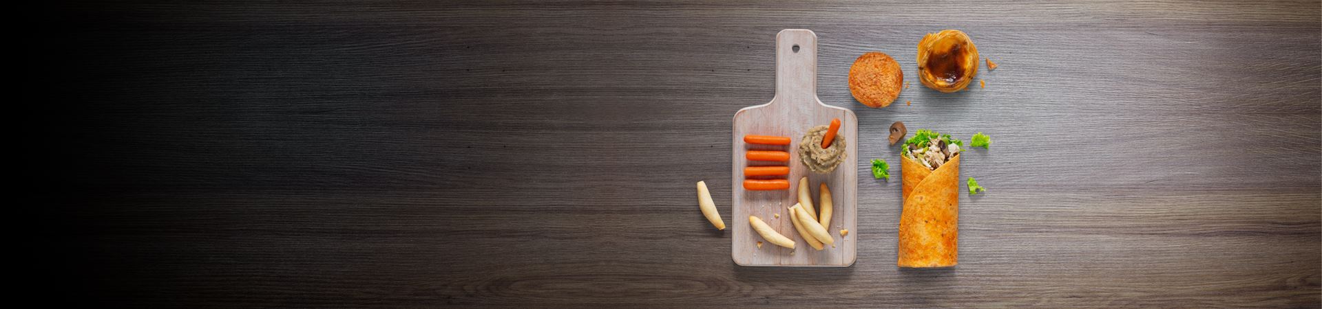 Fotografia de uma mesa de madeira com 4 elementos: um tabuleiro com cenouras, palitos de pão e uma tigela com molho; um wrap de frango, cogumelos e alface; um pastel de feijão e um pastel de nata. 