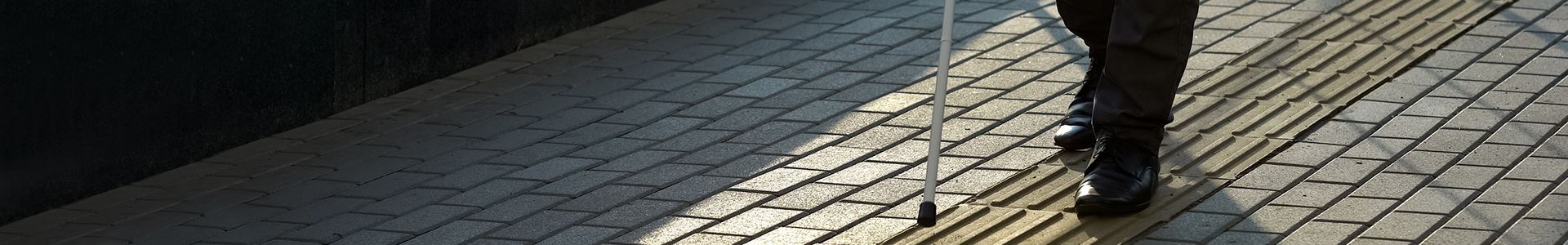 Imagem da parte inferior de um corpo, usando calças e sapatos pretos, e com uma bengala para deficientes visuais, numa calçada cinza com uma barra de piso tátil para deficientes visuais.