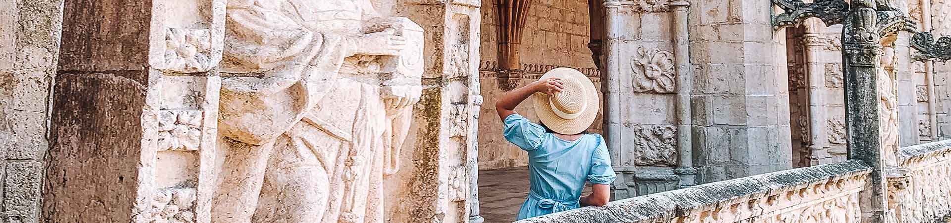 Imagem constituída por uma senhora de costas, a contemplar um monumento de pedra branca. A senhora tem cabelo preto comprido e está com um vestido azul e um chapéu de palha beje que segura com a sua mão esquerda. 