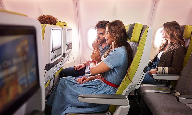 Изображение включает в себя трех пассажиров, сидящих на зеленых сиденьях самолета TAP. Два пассажира смотрят на экраны на спинках передних сидений. Пассажир заднего ряда смотрит в иллюминатор самолета.