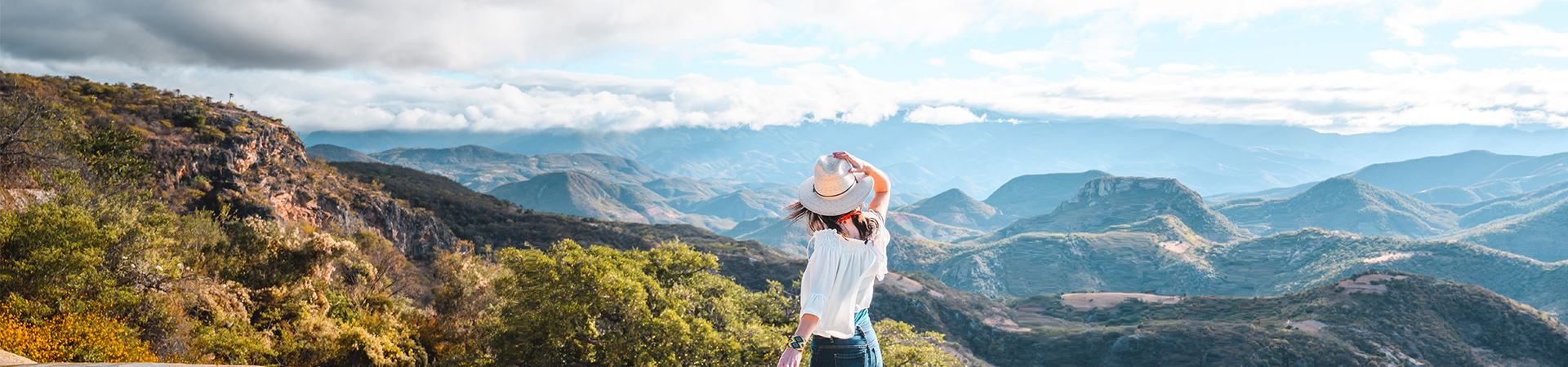 Landscapes of vast mountainous area, with clouds hanging over the top of the mountains. In the foreground you can see a girl, with her back towards the objective, watching the landscape. Her right hand is holding a beige hat with a wide brim. The brown hair is loose and shoulder-length. She wears a white, half-sleeve blouse and blue jeans. On her left wrist she wears a wide, colorful bracelet.
