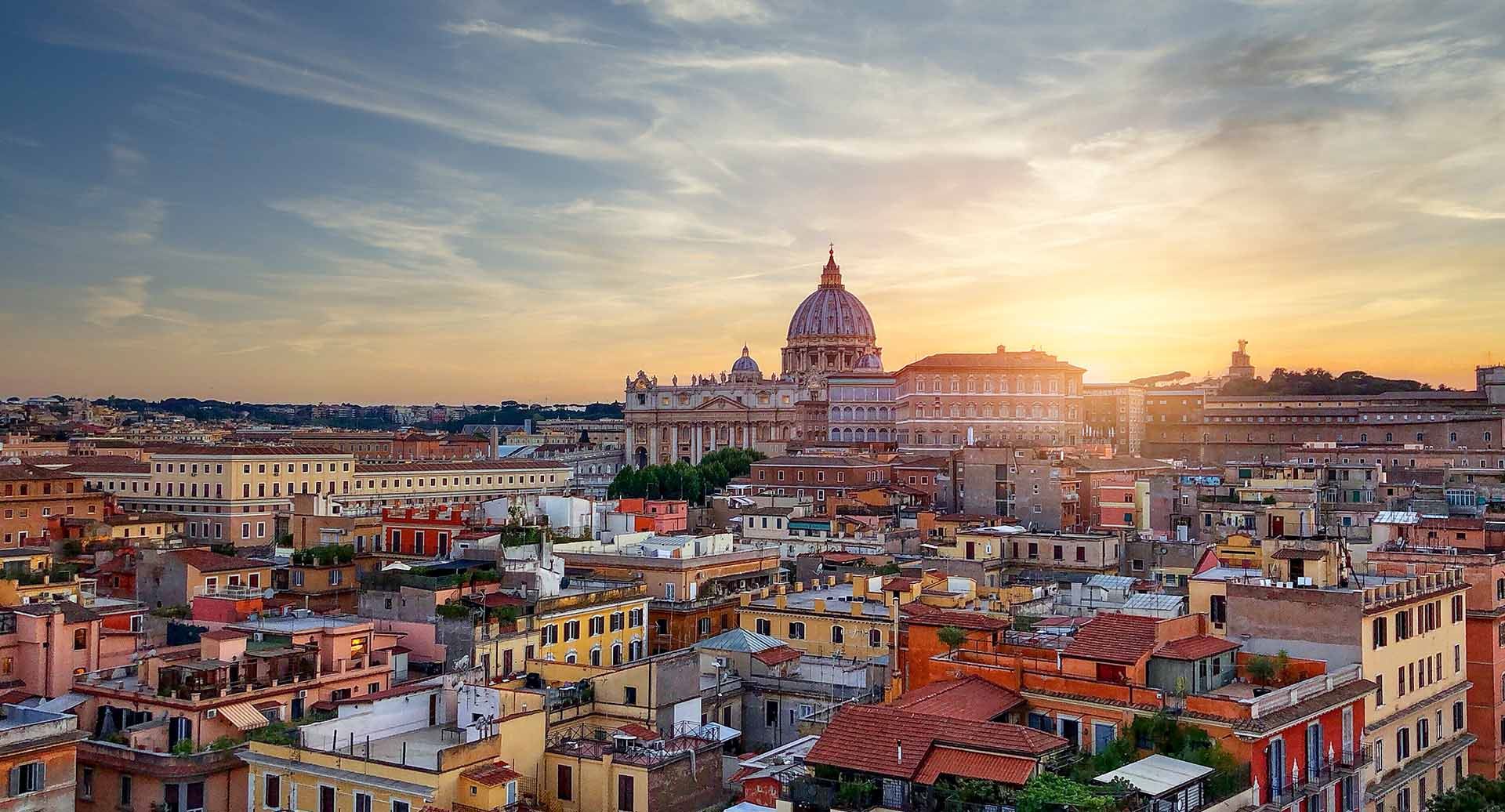 Imagem de um plano aéreo da cidade de Roma. Em primeiro plano, há vários edifícios residenciais, enquanto o plano de fundo mostra vários monumentos romanos e o pôr do sol atrás deles.