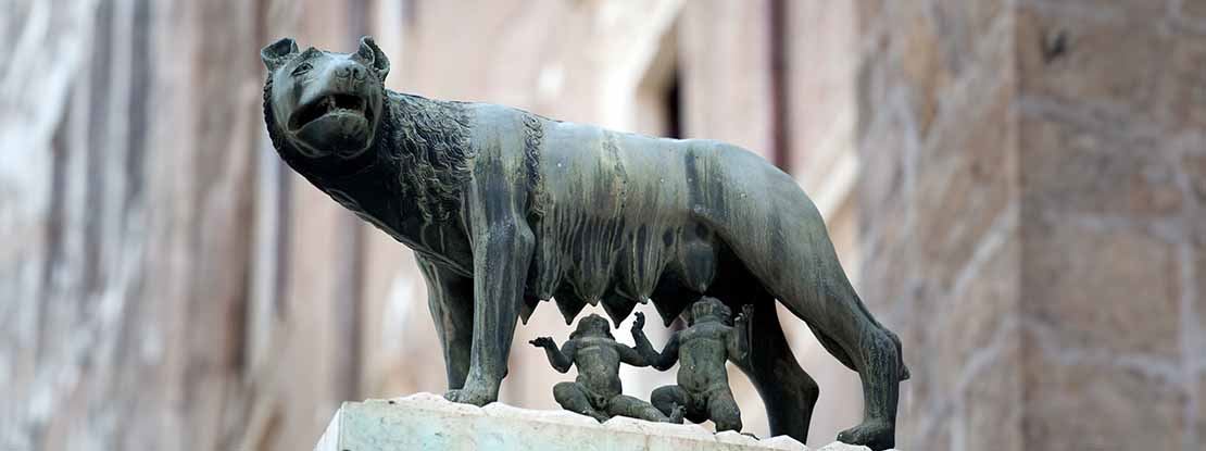 Imagem de ângulo baixo de uma escultura de bronze que representa uma loba em pé em um púlpito de pedra branca enquanto amamenta duas crianças nuas que estão sentadas abaixo dela. Ao fundo, há um edifício romano de pedra bege.