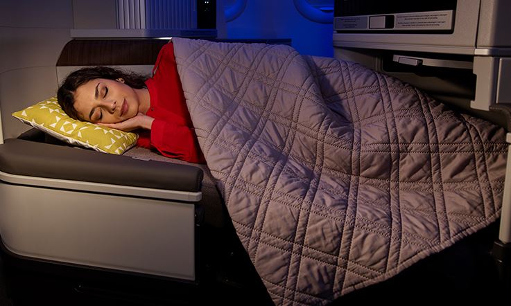 Lying on a reclined airplane seat, which is opened as a bed, a brunette lady sleeps with her head on a pillow. She is covered up to her shoulders with a duvet and has her right arm under her head.