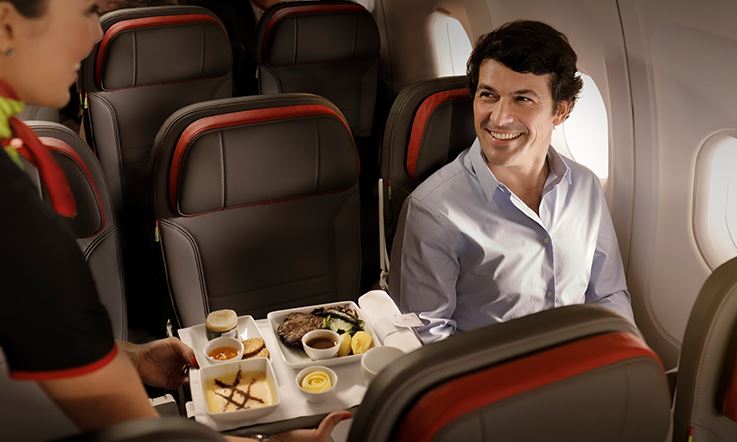 名男子坐在机内靠窗位置的照片。该男子正面带微笑注视着图像左侧的一名空姐，空姐手拿托盘，上面放着几盘食物。