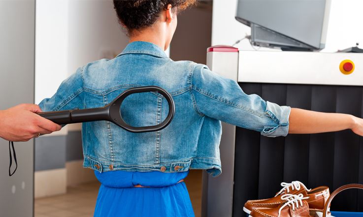 Photo of an open armed woman, facing backward, dressed in a denim jacket, at the airport security checkpoint. On the left side, there is a hand holding a metal detector and leaning it against the woman's back.
