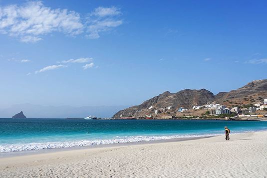 Landscape view of Laginha Beach, where the sand is visible in the foreground, flanked on the left by the sea with two or three shades of blue, and a landscape of hills in the background, where the facade of some buildings can be seen.