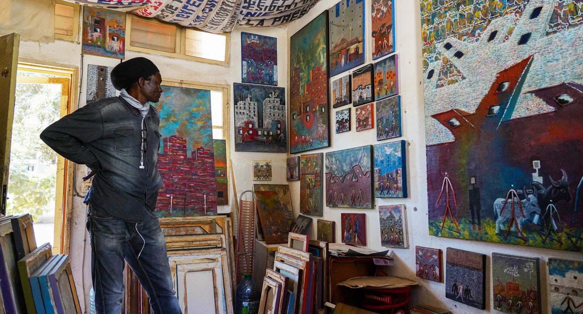 Full-body photo of a man, seen from the side, contemplating a wall that is almost entirely covered with canvases of different sizes, painted in various colors, motifs and styles.