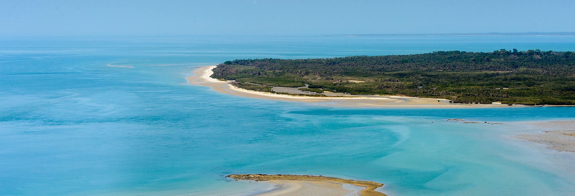 Image of the sea in various shades of blue, with a strip of sand and vegetation visible in the background, extending from the right side to the center of the photograph.