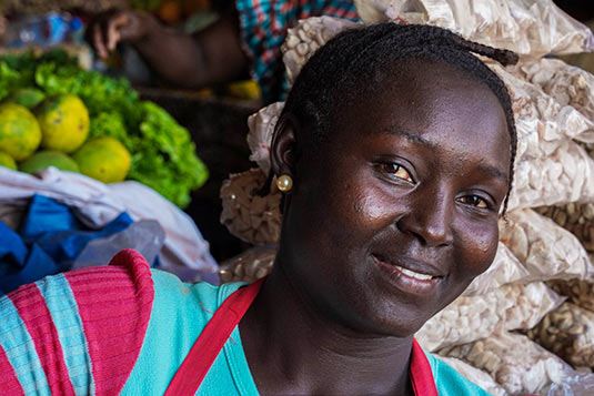Photograph of a smiling Cape Verdean woman from the chest up. The woman is wearing a t-shirt and apron and has a series of fruits and vegetables and piled up sacks behind her, suggesting that she could be a market vendor.