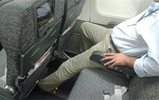 Photographie d'un homme assis à l'intérieur d'un avion, à côté de la fenêtre dans une rangée de deux sièges, et avec sa jambe gauche allongée sous le siège d'avant.
