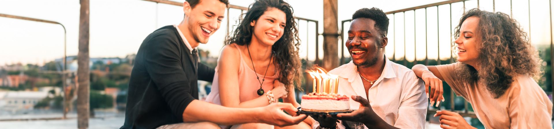 Esta fotografía muestra a cuatro personas (dos chicas y dos chicos) sonriendo y sentadas al aire libre. Los dos chicos sostienen una tarta de cumpleaños con varias velas encendidas.