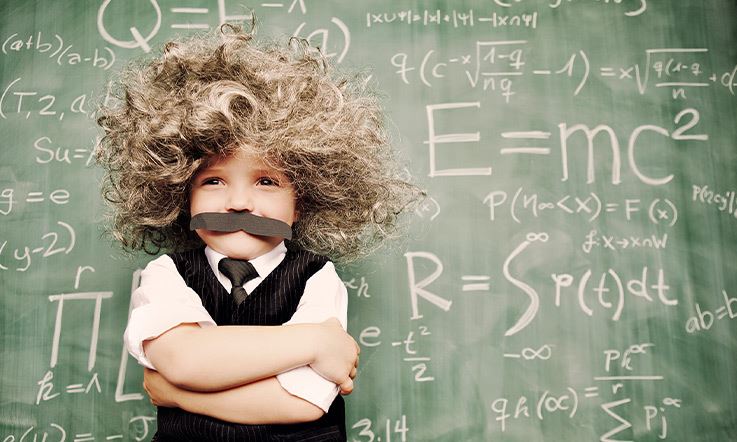 A smiling boy wearing a suit, a tie, a cardboard mustache, and a wig with a lot of hair, recalling the brilliant Albert Einstein. Behind him stands a green slate board with several equations written in chalk.