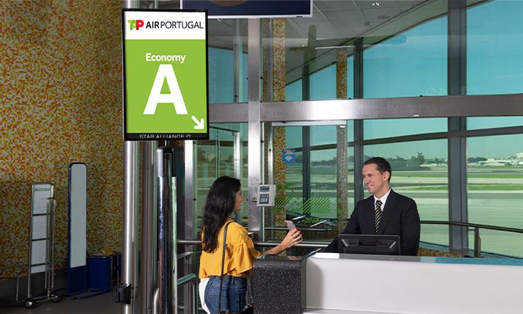 Image of a service desk in the boarding area. A gentleman in a dark suit is attending a lady wearing a yellow blouse and jeans. The lady holds the cell phone with her right hand and is showing some information. Next to the desk there is a sign with the TAP Air Portugal logo on it and, below the logo, the word “Economy” followed by a large size letter “A” highlighted in a green background.