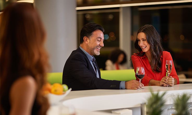 Image of a gentleman and a lady, caucasian, with dark hair, sitting at the bar of a TAP Lounge. They are facing the image with the white bar counter in front of them, on top of which are two glasses of red wine that they both hold in their hands. In the foreground of the image, on the left, there is a lady facing the background, with long brown hair.