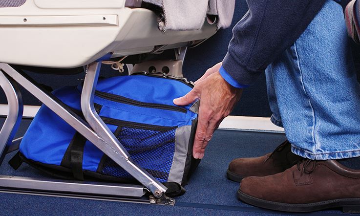 As pernas e os braços de um passageiro, sentado num assento de avião, num ambiente iluminado. Está a arrumar uma mochila azul debaixo do assento da frente.