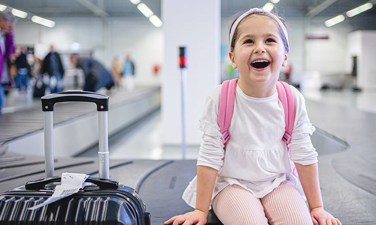 A smiling child, with a hold suitcase next to her, sits at the foot of the baggage collection treadmill, inside an airport. The room is lit up, and in the background there are several people collecting their belongings.