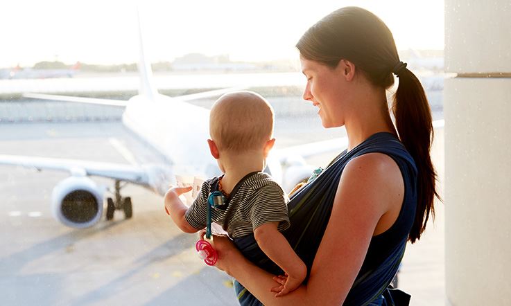 A woman, facing laterally, is standing, and holds a baby on her lap, with her back to the image. They are inside an airport and watch from the window an airplane on the runway, illuminated by daylight.