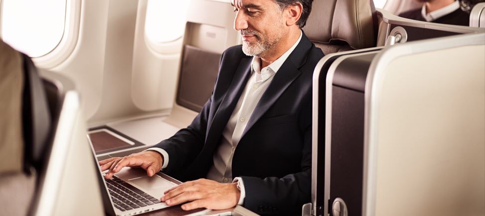 Close-up frontal shot of a gentleman sitting on the Executive Class seats. He has his laptop on the folding table and he is writing on it. The gentleman is dark, with a gray beard, and wears a black blazer and a white shirt.
