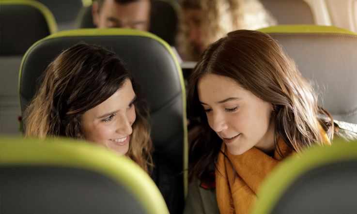Close-up of two ladies, visible up to their shoulders, in standard seats. They are brunettes and they are sitting side by side, smiling. The one on the left is looking at her colleague and the one on the right is looking down.