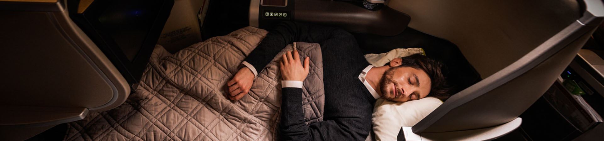 Low light environment. We see a young man lying down, eyes closed, on an Executive Class bed. His head is on the headboard and is covered with a gray duvet. Above his legs hangs, suspended, a TV screen.