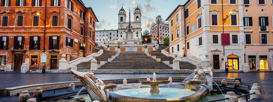 Image d'une rue de Rome sur laquelle, au premier plan, on voit une fontaine et, en arrière-plan, un escalier en pierre blanche, flanqué de deux bâtiments, tous deux dans les tons orange. Au bas de l'escalier se trouve un bâtiment en pierre blanche qui ressemble à une église à deux tours.