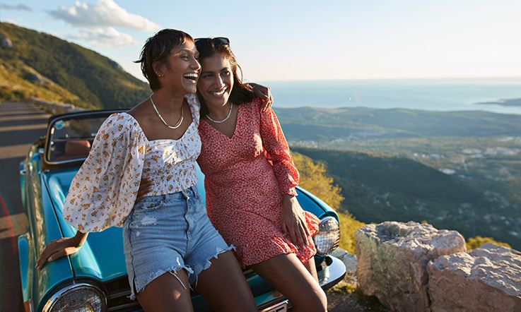 Photograph of two brunette women laughing and leaning against the front of a blue car parked at a scenic viewpoint. The woman on the left is wearing denim shorts and a white blouse with flowers. The woman on the right is wearing a pink dress. Behind them is a landscape with mountains and the sea in the background. 