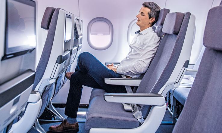 Photograph of a man sitting alone in an airplane in a row of 3 chairs. The man is sitting in the center of the row of chairs with his left leg resting on top of his right leg. He is wearing blue pants and a white shirt. 
