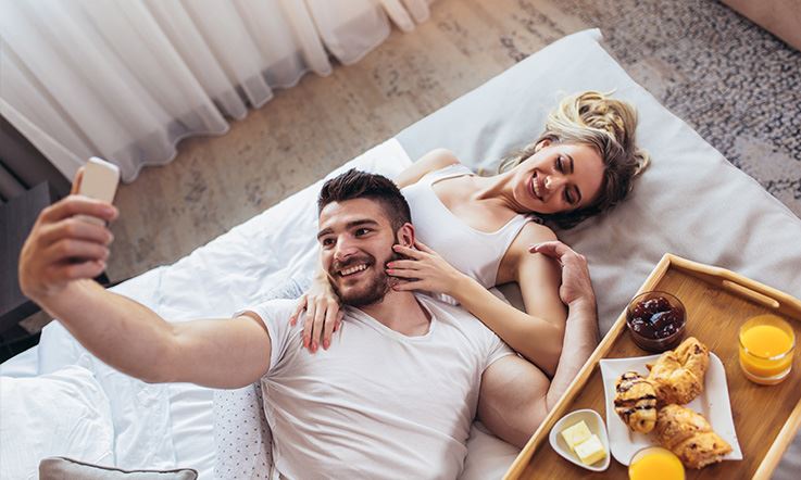 Photograph of a man and a woman lying on a bed in a hotel room. The man has his head resting on the woman's belly and is holding his cell phone as if taking a selfie of them. Next to them is a tray with croissants, orange juice, butter and jam. 