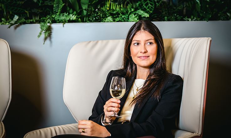 Foto einer brünetten Frau, die mit einem Glas Wein in der Hand in einem Sessel einer Lounge sitzt. Die Frau trägt einen schwarzen Blazer über einem weißen Hemd. 