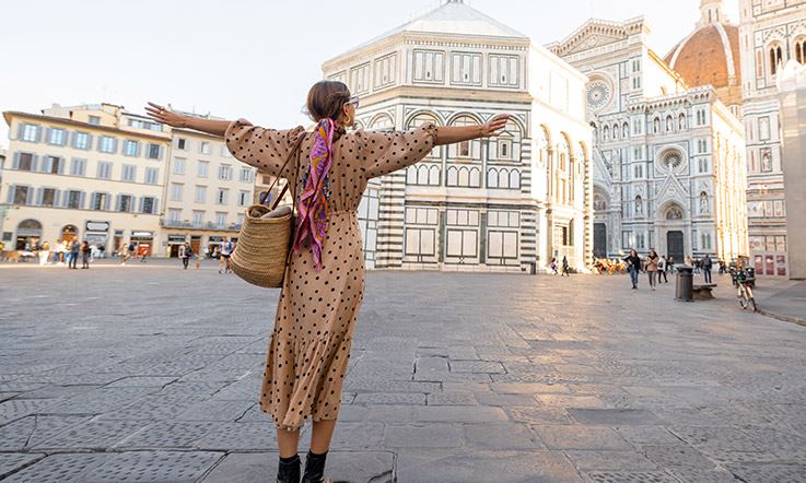 Photograph of a woman with open arms in the cathedral square in Florence. The woman is facing the cathedral and is wearing a long dress with flowers. 