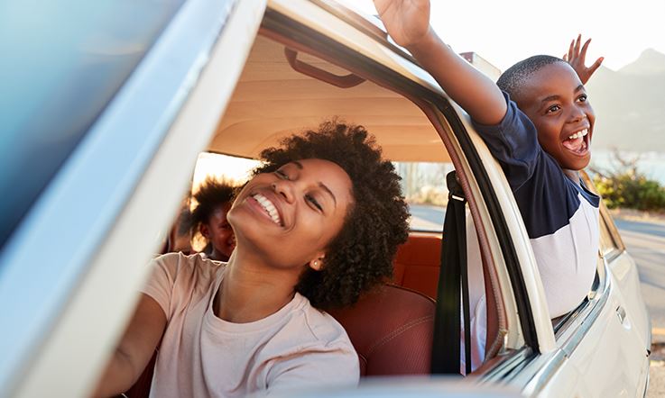 Foto einer Frau und zweier Kinder in einem weißen Auto mit geöffneten Fenstern. Die Frau lächelt auf dem Fahrersitz. Auf dem Rücksitz lehnt sich eines der Kinder mit ausgestreckten Armen aus dem Fenster und lächelt. 