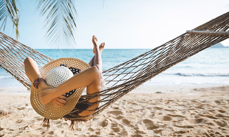 Photograph of a woman lying in a hammock on a beach. The woman is facing the sea and has a large hat, which she holds with both hands, while her feet are crossed in the air. 