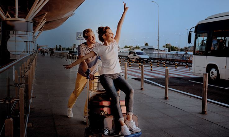 Photograph of a man and a woman outside an airport. The man is pushing a trolley with suitcases, while the woman is sitting in front of the trolley with her arms open. They are on the sidewalk next to the airport, with the road on the right side where a bus is stopped next to a pedestrian crossing.