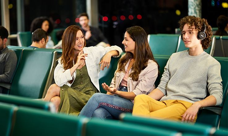 Three people – two women and a boy – are sitting on chairs at the airport. The two women talk excitedly. Next to them, there's a quiet boy wearing headphones. In a blurred shot, other people can be seen sitting. 