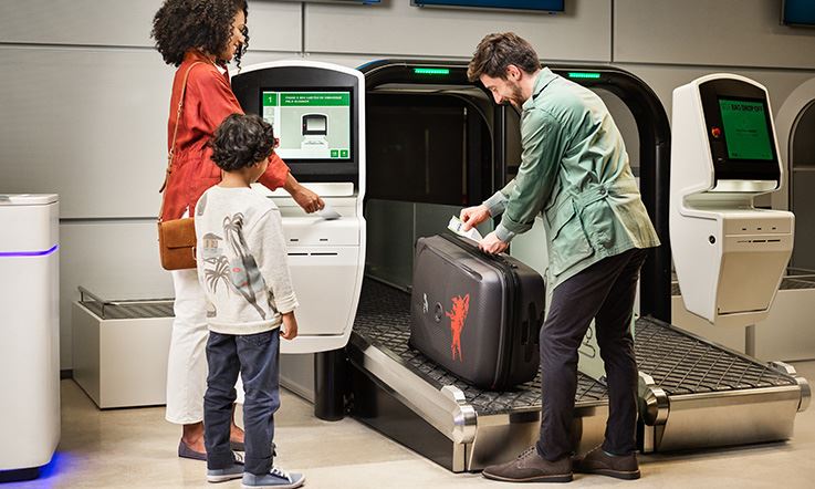 Three passengers – a couple and a boy – are standing at the airport, next to a counter to check in their luggage. The man grabs a luggage tag that is attached to the hold suitcase. The suitcase is on a conveyor belt to be weighed and checked. The woman is holding a ticket. She and the boy are waiting next to the man. 